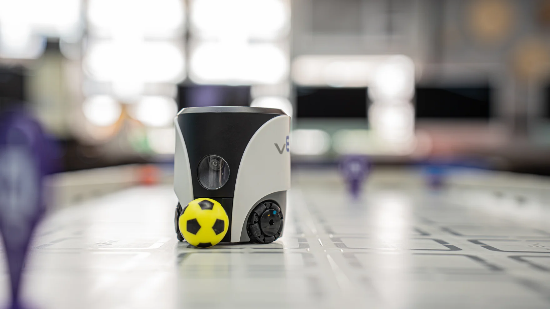 A VEX AIM robot beside a yellow ball on a robotics activity mat